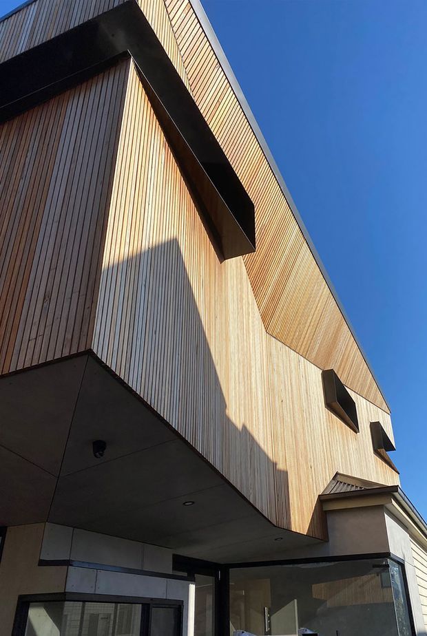 Upward-looking exterior view of a contemporary multi-level building facade clad in closely spaced vertical timber boards. Bold black projecting geometric eaves or balcony forms create strong shadows across the warm wood surface, with dark-framed glazing and a covered entry visible at the lower level against a clear blue sky.
