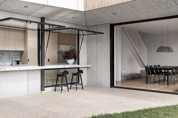 Contemporary indoor-outdoor entertaining area with a concrete kitchen and bar opening onto a dining room. The space features pale timber cabinetry, a light stone benchtop, black steel framing, exposed concrete ceilings and walls, and large glazed sliding doors. The adjacent dining area contains a long dark dining table, black chairs, and pendant lights. A small strip of lawn is visible at the threshold.