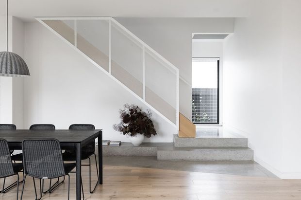 Bright minimalist open-plan dining and stair area with a black rectangular dining table and woven chairs in the foreground. A floating or partially enclosed staircase with white balustrade panels rises along the wall, transitioning to a warm timber stair section and broad concrete steps. Pale timber flooring, white walls, a slim black-framed window, recessed ceiling detailing, and a sculptural pendant create a clean, contemporary interior.