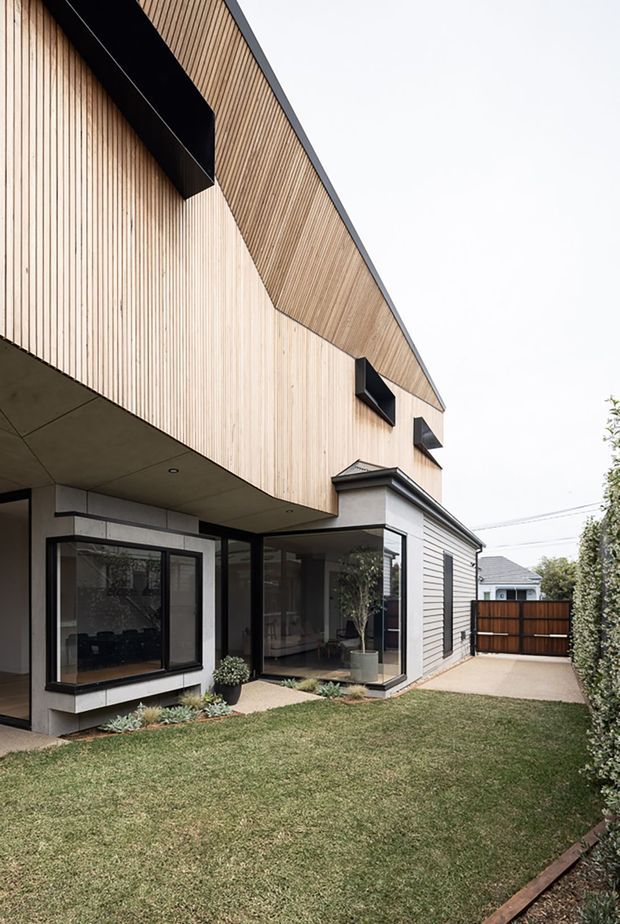 Contemporary two-storey residential exterior with a sharply angled roofline and upper facade clad in narrow vertical light timber battens. The lower level combines pale horizontal cladding, dark-framed floor-to-ceiling glazing, and a deep concrete soffit, opening onto a small landscaped lawn and paved side path. Black projecting awnings and trims provide strong contrast, with a timber privacy gate and neighbouring homes visible in the background.