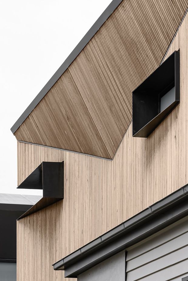 Close-up exterior architectural photograph of a contemporary gabled building clad in vertically ribbed light timber, with sharply projecting black-framed window boxes and dark metal roof edges. A lower section with horizontal light cladding and prominent guttering is visible in the foreground.