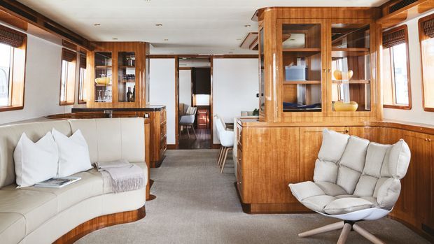 A bright, refined interior lounge and dining area with extensive warm timber cabinetry and built-in display shelving, cream upholstered seating, carpeted floors, and multiple windows. The open-plan space extends toward a dining area and hallway, with a strong mid-century-inspired built-in joinery aesthetic.