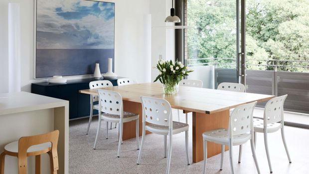 Bright contemporary dining area with a large light timber dining table, white perforated dining chairs, and a linear pendant light. The room has white walls, grey carpet, expansive glazing opening toward leafy greenery, and a dark sideboard beneath a large coastal-toned artwork.