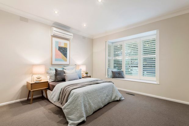 A bright, neatly styled bedroom with a low-profile bed dressed in layered pale blue and grey bedding, matching bedside tables and lamps, soft neutral walls, recessed ceiling lights, carpeted flooring, framed artwork, and a large window fitted with white plantation shutters. A wall-mounted split-system air conditioner is positioned above the artwork.
