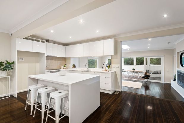 Bright open-plan contemporary kitchen with extensive glossy white cabinetry, a large white island with four stools, pale benchtops and splashback, and dark polished timber flooring. The kitchen opens to a dining or living area with full-height glazing, while a wall-mounted fireplace is visible at the right.