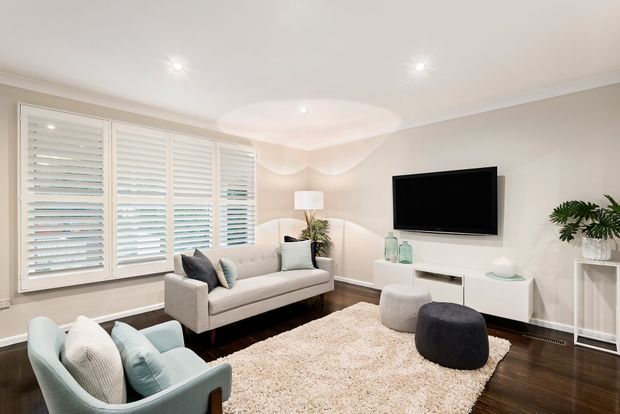 Bright, compact living room with a neutral upholstered sofa, pale blue armchairs, a wall-mounted television, white low-profile media cabinetry, textured area rug, indoor plants, and dark timber flooring. Large plantation shutters cover the windows, while recessed ceiling lights and decorative ceiling detailing create a polished, contemporary interior.