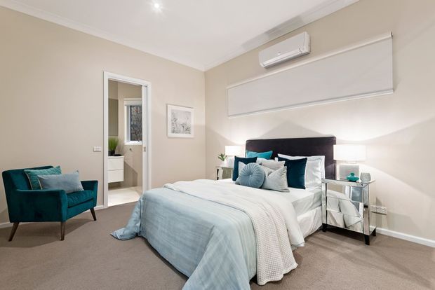 A bright, refined bedroom with a neatly made bed, upholstered dark headboard, teal accent cushions and armchair, mirrored bedside tables, beige walls, carpeted flooring, and a roller blind. An open doorway reveals a small adjoining bathroom or dressing area.