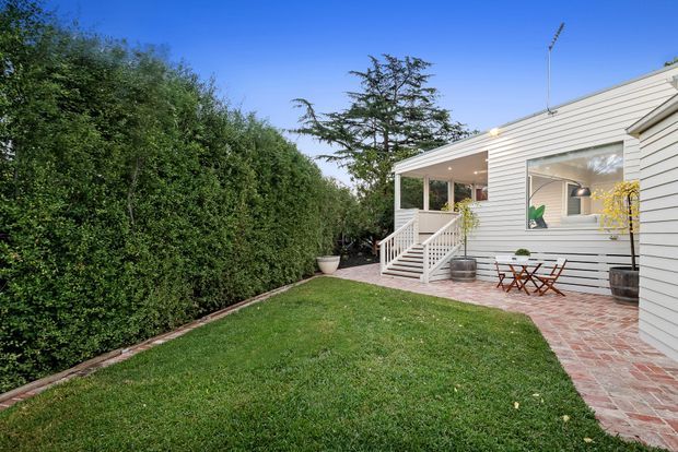 Exterior view of a single-storey suburban home with pale horizontal weatherboard cladding, a small covered porch and stair entry, and a brick-paved patio with outdoor bistro seating. A neatly trimmed hedge borders the lawn and creates a private garden setting.