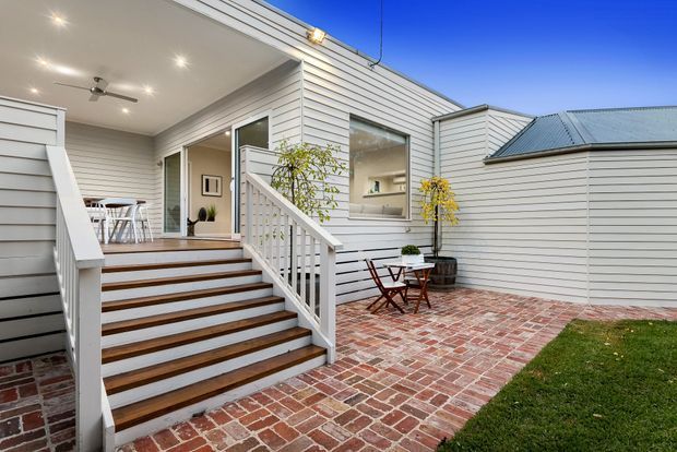 A bright residential courtyard and raised deck entrance with white horizontal weatherboard cladding, a low metal gabled roof, broad timber steps with white railings, and an outdoor dining setting. Red brick paving defines the courtyard, with a small lawn and potted greenery adding softness. The open glazed doorway reveals a dining area inside.