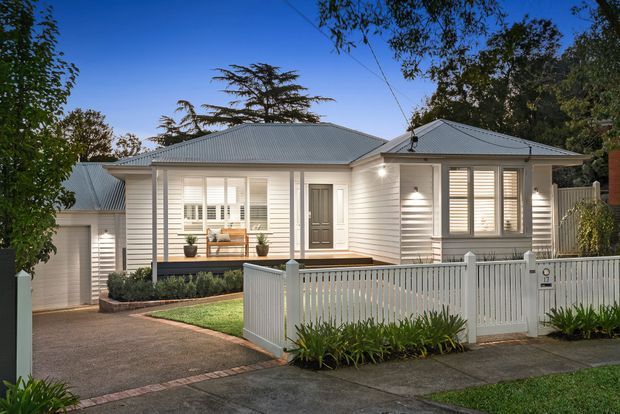 Street-facing view of a single-storey suburban home with a light weatherboard facade, multiple grey corrugated metal pitched roof sections, white-framed windows with shutters, a small covered front porch, enclosed front garden, driveway, and attached garage. The house has a refined traditional bungalow-like appearance.