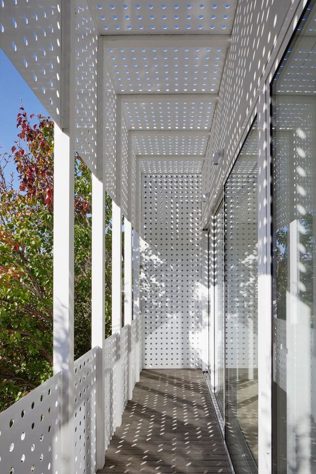 A narrow, covered exterior balcony or walkway beside a glazed building facade. White perforated architectural screens form the balustrade, ceiling, side wall, and overhead canopy, casting a repeating pattern of light and shadow across the grey floor and reflective glazing. Slender white columns frame views toward leafy trees and blue sky, creating a bright, contemporary screened outdoor passage.