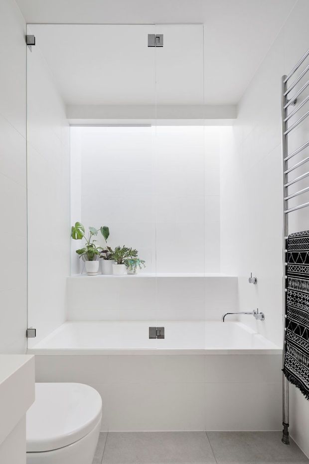 Bright, minimalist white bathroom with a built-in bathtub beneath a large mirrored or glass-panelled wall and recessed ledge displaying small potted plants. A modern chrome towel rail with a black-and-white patterned towel is visible at right, with a white toilet in the foreground. The room has pale grey floor tiles, white tiled walls, clean rectangular detailing, and abundant diffuse light.