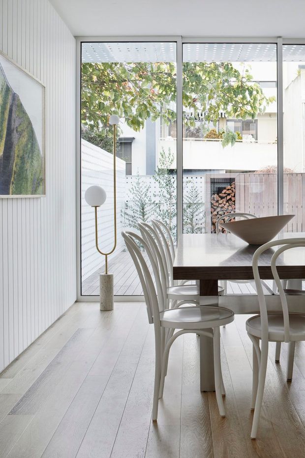 Bright, minimalist dining room with a white timber dining table and curved white chairs positioned beside full-height glazed doors. Pale timber flooring and vertically panelled white walls create a clean, light interior. Through the glazing is a small courtyard with timber decking, layered planting, a privacy screen, and a built-in firewood niche. A sculptural floor lamp and large framed artwork add restrained decorative elements.