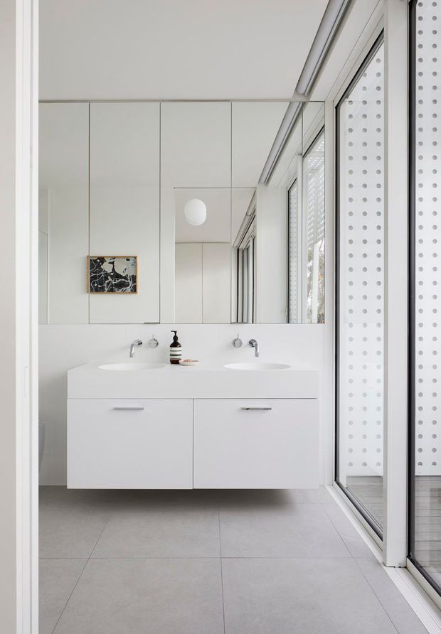 A bright, minimalist bathroom with a floating white double vanity, two integrated basins, wall-mounted chrome taps, mirrored cabinetry, and large light-grey floor tiles. Full-height glazing lines one side of the room, accompanied by a white perforated privacy screen. The space has clean linear detailing, abundant natural light, and restrained contemporary styling.