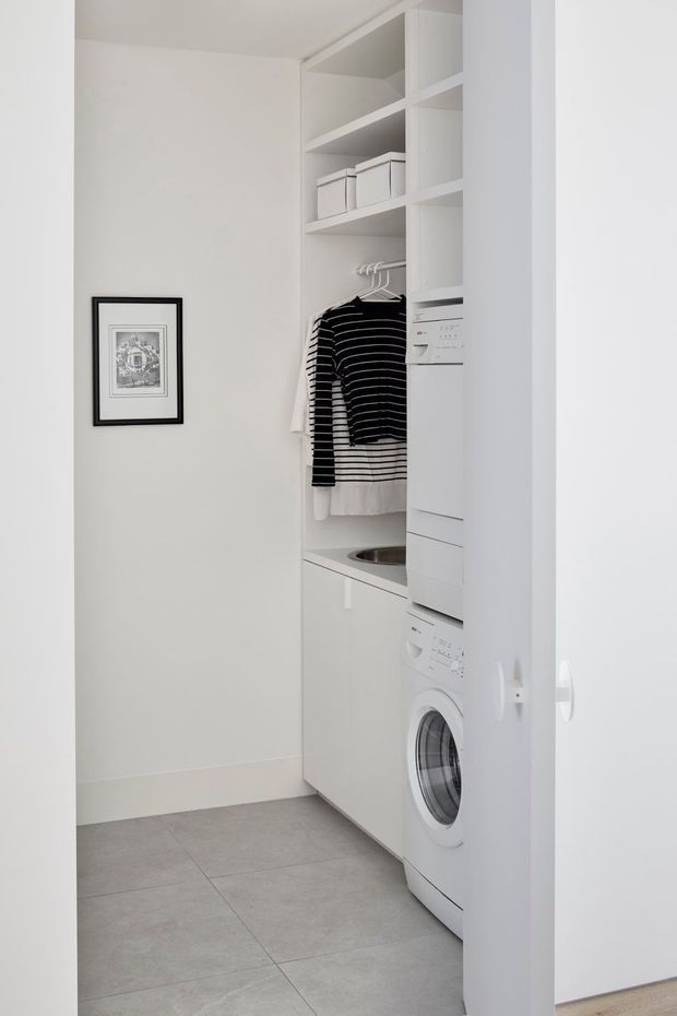 A compact, bright laundry room with white built-in cabinetry, open shelving, hanging rails, a front-loading washing machine, and an integrated utility sink. The room has pale grey large-format tiled flooring, white walls, and minimal black-and-white wall art.