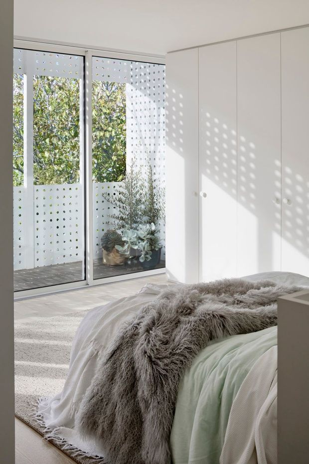 Bright, minimalist bedroom with a partially visible bed dressed in white and pale green bedding and a large shaggy grey throw. Full-height white built-in wardrobes occupy the right wall. Floor-to-ceiling glazed doors open to a small balcony or exterior space with perforated privacy screening, casting a dotted pattern of sunlight across the room. Potted plants are visible outside. The space has a calm, airy, neutral palette and abundant natural light.