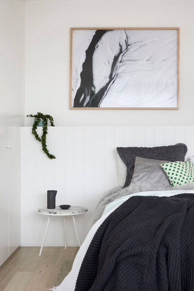 A bright, compact bedroom with a white vertical-panel feature wall, pale timber flooring, and a partially visible bed dressed in layered grey, black, white, and green textiles. A small round side table with a dark vase and bowl sits beside the bed, while a trailing plant and large monochrome abstract artwork add decorative accents. The overall look is clean, contemporary, and softly layered.