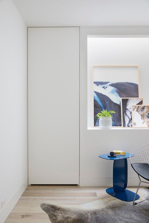 A bright, minimalist corner of a contemporary interior featuring a tall flush white built-in door or wardrobe, pale timber flooring, and a recessed display niche with framed abstract artwork and a potted fern. A small sculptural blue side table, coffee cup, books, wire-frame chair, and neutral hide rug add restrained colour and texture.