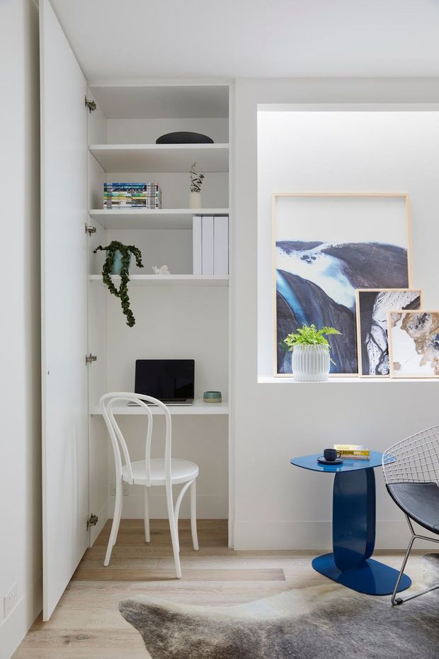 Bright, compact home office nook integrated into a white built-in shelving unit, with a small desk, laptop, white chair, books, and decorative objects. The adjacent room includes framed artwork, a blue side table, a wire-frame chair, and a neutral hide-style rug. The space has a clean, contemporary appearance with pale timber flooring and white joinery.