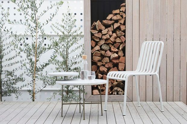 A styled outdoor seating area with a white slatted chair and small round tables on a pale timber deck. A vertical stack of firewood is recessed into dark wall cladding, while olive trees stand in front of a light patterned screen. The scene has a restrained Scandinavian-influenced contemporary aesthetic with pale wood, white furniture and natural greenery.