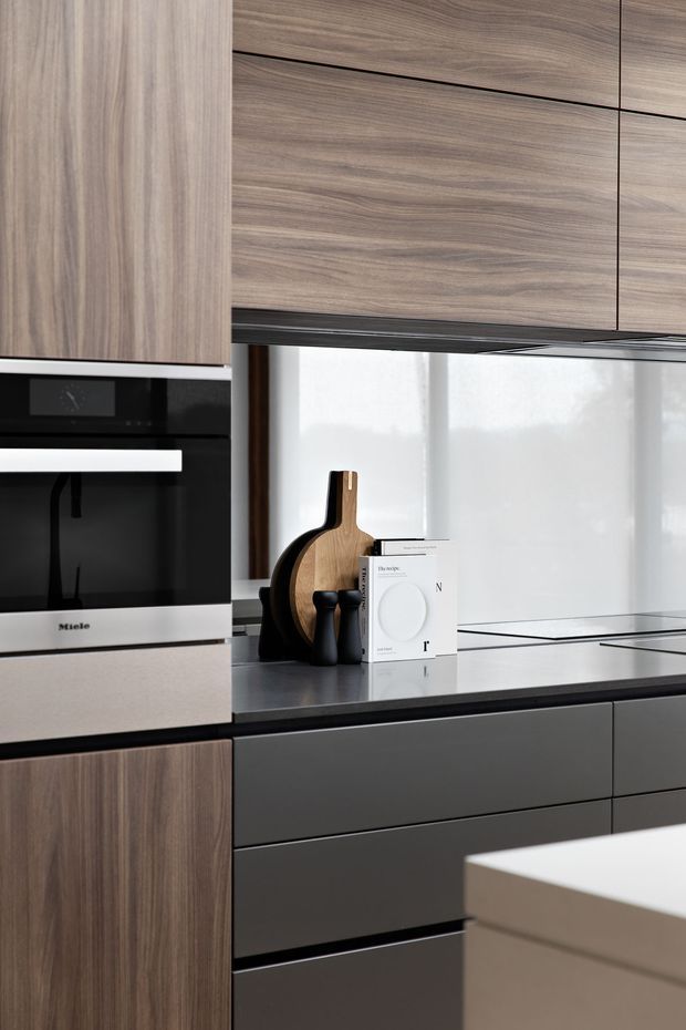 Close-up view of a contemporary kitchen featuring full-height walnut-toned woodgrain cabinetry, dark matte lower drawers, a black induction cooktop, dark benchtops, and an integrated black-and-stainless Miele oven. A light translucent window covering provides soft natural light behind the countertop, while cutting boards and small kitchen accessories sit on the bench.