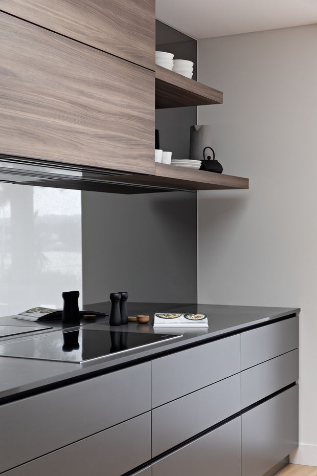 A close-up of a refined contemporary kitchen with dark grey handleless cabinetry, a dark stone benchtop and matching splashback, and warm wood-grain overhead cabinetry with open display shelving. Minimal ceramic tableware and a black kettle sit on the shelves and benchtop. The composition highlights clean horizontal lines, integrated appliances and a restrained neutral palette.