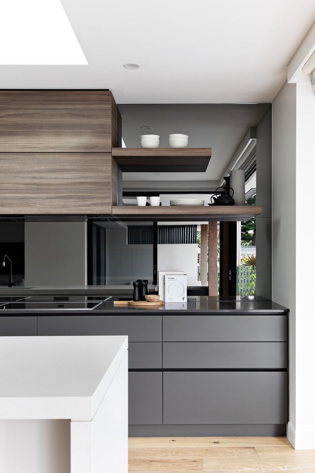 A contemporary galley-style kitchen with dark grey flat-panel cabinetry, a black stone benchtop, integrated cooktop, and open timber shelving. A large white island occupies the foreground, while warm timber flooring and extensive glazing bring natural light into the space. The palette is restrained and sophisticated, with charcoal, white, timber, and metallic accents.