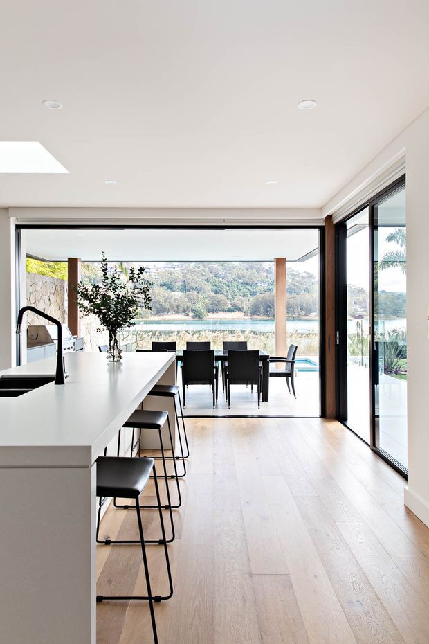 Bright contemporary open-plan kitchen opening to an outdoor dining terrace and pool, with a large white island, black bar stools, pale timber flooring, extensive black-framed glazing, and views toward a landscaped waterfront setting.