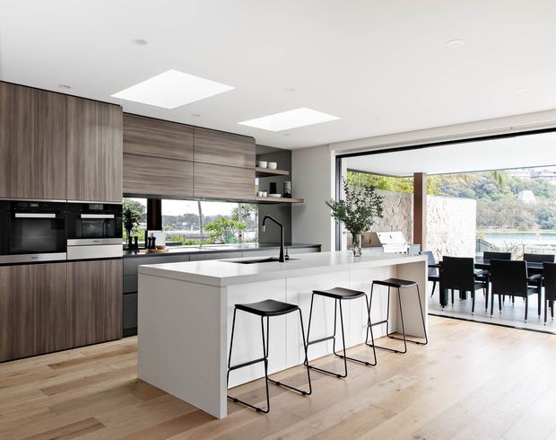 Bright contemporary open-plan kitchen with a large white island, four black bar stools, dark wood-grain cabinetry, integrated appliances, timber flooring, and expansive sliding glass doors opening to a dining and outdoor area with a water view. Two large skylights add natural light.