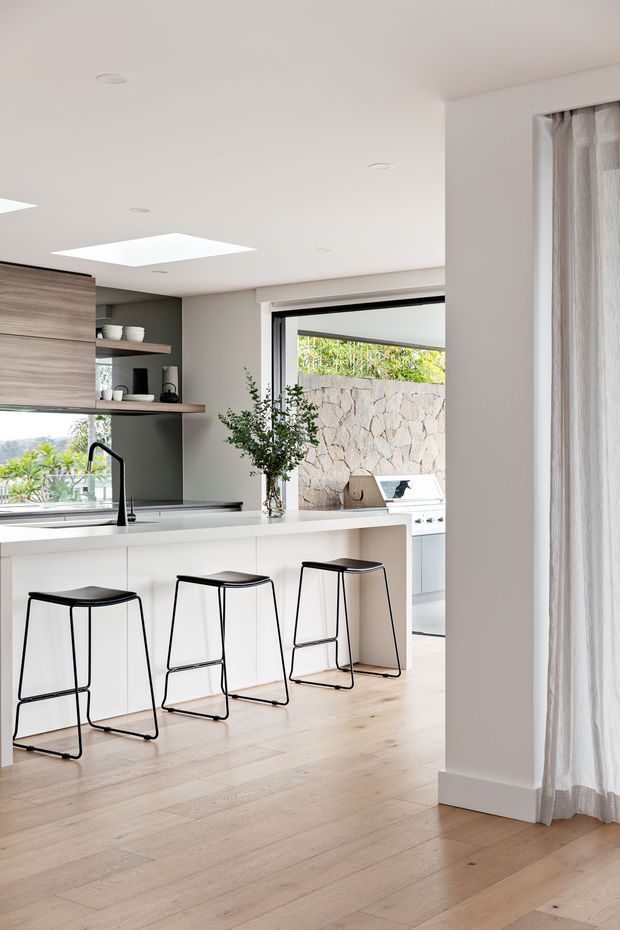 Bright contemporary kitchen with a white island and three black metal bar stools, pale timber flooring, light wood cabinetry, open shelving, and a dark grey-green splashback wall. A large opening leads to an outdoor cooking area with a stone-clad wall and built-in barbecue. Floor-to-ceiling sheer curtains and skylights add abundant natural light.