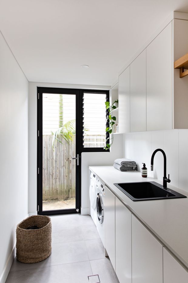 A bright, narrow contemporary laundry room with white full-height cabinetry and a long white benchtop containing a black inset sink and curved black tap. A front-loading washing machine, folded towels, soap dispenser, woven basket, indoor plant, tiled floor, and black-framed glazed exterior door are visible. The space has clean minimalist detailing and strong natural light.