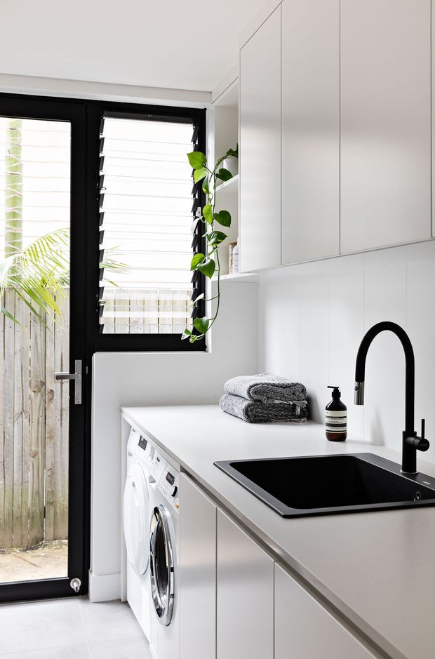 Bright, compact contemporary laundry with white built-in cabinetry and benchtop, integrated front-loading washing machine, black undermount sink and tall black tapware. Black-framed glazed door and louvre window provide ventilation and daylight, while a trailing indoor plant and folded towels add softness.
