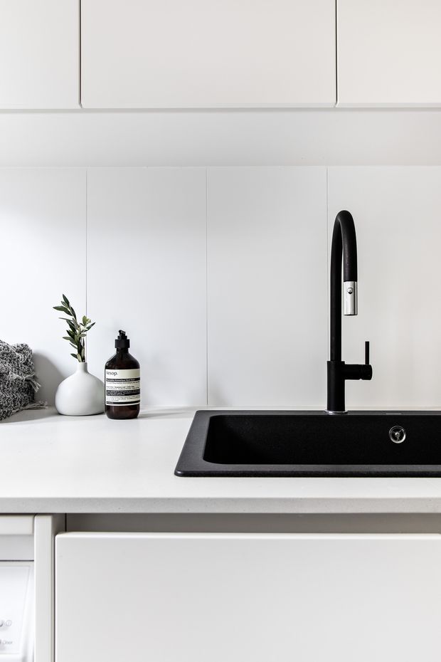 Close-up of a clean, minimalist kitchen sink area. White flat-panel cabinetry and a white benchtop surround a large black inset sink with a tall black-and-chrome gooseneck tap. A small white vase with greenery and a dark soap dispenser sit on the counter.