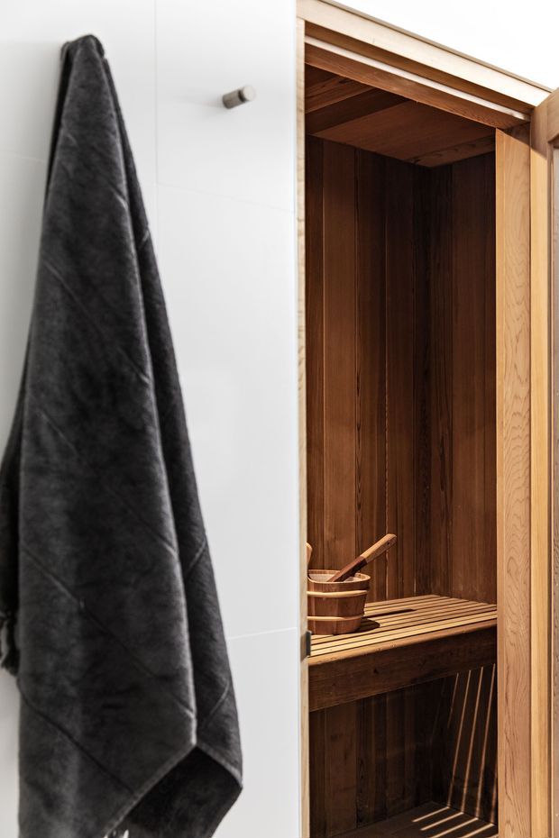 Close-up interior view of a compact timber-lined sauna enclosure within a white-tiled room. The sauna has dark vertical wood wall cladding, a slatted timber bench, a wooden bucket and ladle, and a natural timber-framed door. A dark towel hangs on the adjacent wall.