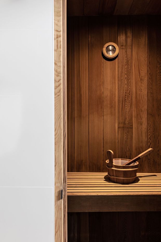 Close-up view of a compact timber-lined sauna interior, featuring vertical dark wood wall and ceiling boards, a slatted wooden bench, and a wooden bucket with ladle. A circular sauna light or vent is mounted high on the wall. The scene has warm, low lighting and rich natural wood tones.