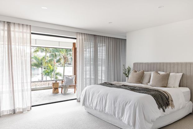Bright, refined bedroom with a large upholstered channel-tufted bedhead, layered white bedding, neutral cushions, and a soft grey throw. Full-height sheer curtains frame expansive sliding glazing that opens to a balcony or terrace with tropical greenery beyond. The room has a simple recessed-light ceiling, pale carpet, and a calm neutral palette.