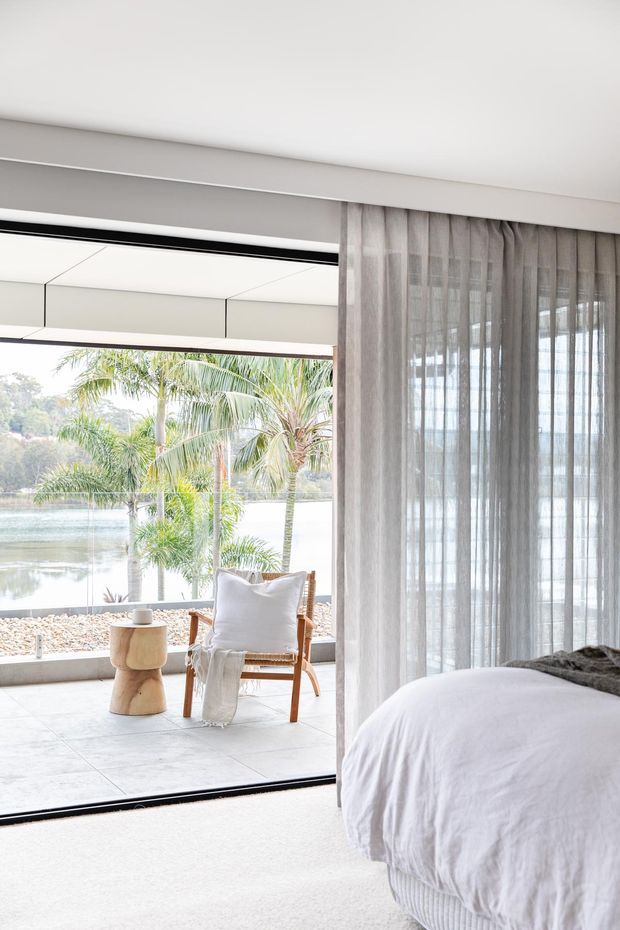 Bright, serene bedroom opening onto a waterfront-facing terrace through large sliding glass doors. The room features sheer floor-to-ceiling curtains, a white upholstered bed with textured bedding, pale flooring, and a light timber lounge chair with a small sculptural timber side table outside. Tropical palms and water are visible beyond the glazing.