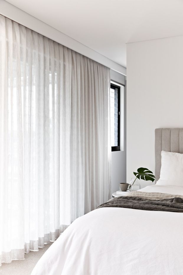 Bright, serene bedroom with a predominantly white and soft neutral palette. Floor-to-ceiling sheer curtains cover large glazing along one wall, while a simple upholstered bed with a vertically channelled beige headboard occupies the foreground. A small round bedside table holds a cup and a potted tropical plant, and layered neutral bedding adds subtle texture.