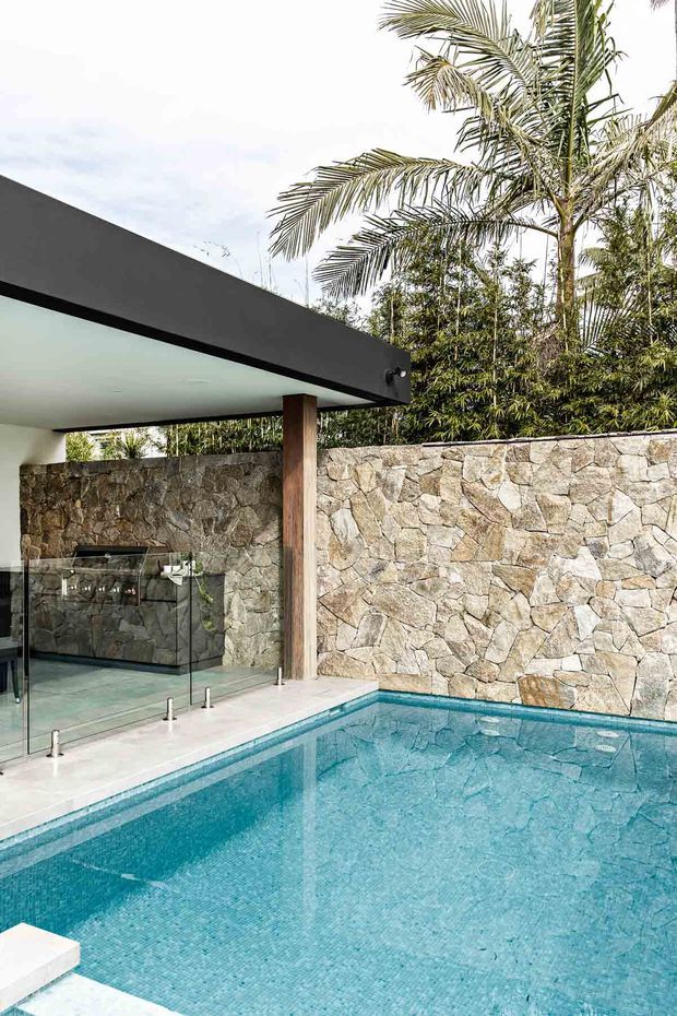 An outdoor pool area beside a covered pavilion with a compact outdoor kitchen or bar behind frameless glass balustrading. The pool is finished in small blue mosaic tiles, while a rough natural-stone boundary wall and tropical planting create a private resort-like setting. The pavilion has a flat dark roof, pale ceiling, and timber support post.