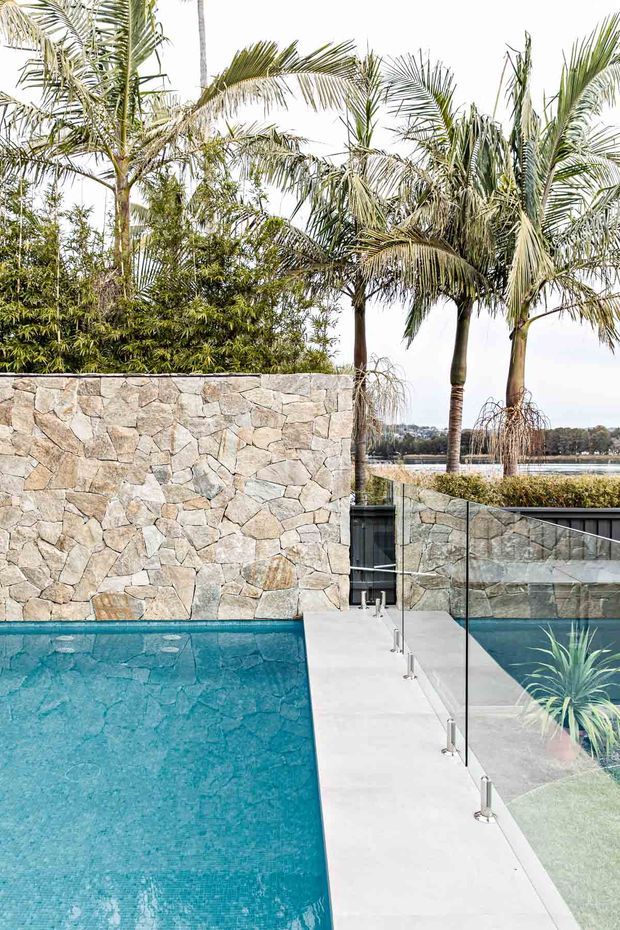 Contemporary outdoor pool area with a turquoise swimming pool, pale concrete pool deck, frameless glass balustrade, and a textured irregular stone-clad privacy wall. Tropical palms and dense planting surround the pool, with a calm body of water visible beyond the property.