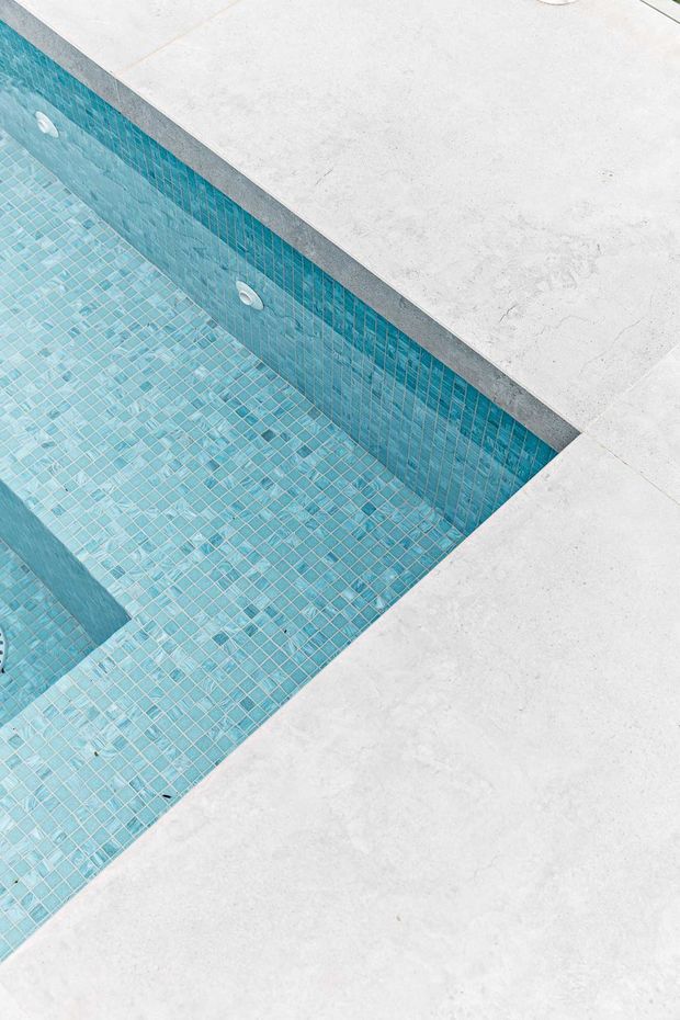 Close-up overhead view of a rectangular swimming pool with pale turquoise mosaic tiles lining the basin and walls, bordered by a broad light grey-white concrete or stone coping and surrounding terrace.