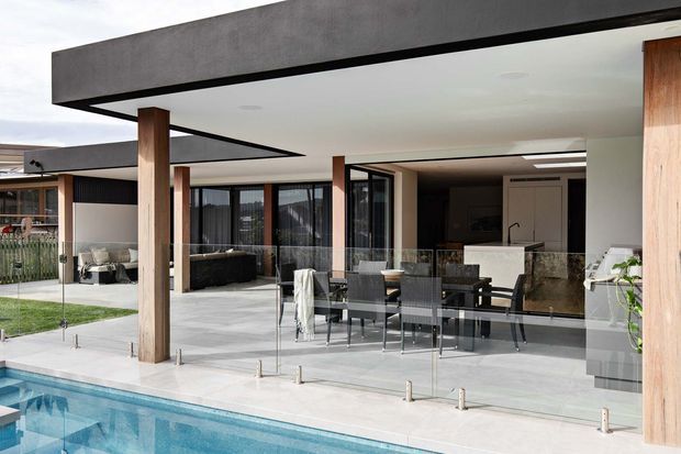 Contemporary single-storey home viewed from the pool, featuring a broad flat roof with deep cantilevered overhangs, timber-clad structural posts, extensive glazing, a covered outdoor dining terrace, and an open-plan kitchen and living area. A glass balustrade separates the terrace from the swimming pool, creating a strong indoor-outdoor connection.