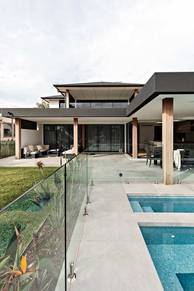 Contemporary two-storey residence with broad cantilevered rooflines, covered outdoor living areas, timber-clad structural columns, dark framed glazing, and a light paved terrace surrounding a tiled swimming pool. A transparent glass balustrade separates the pool terrace from landscaped garden beds, while indoor and outdoor seating areas are visible beneath the covered sections.