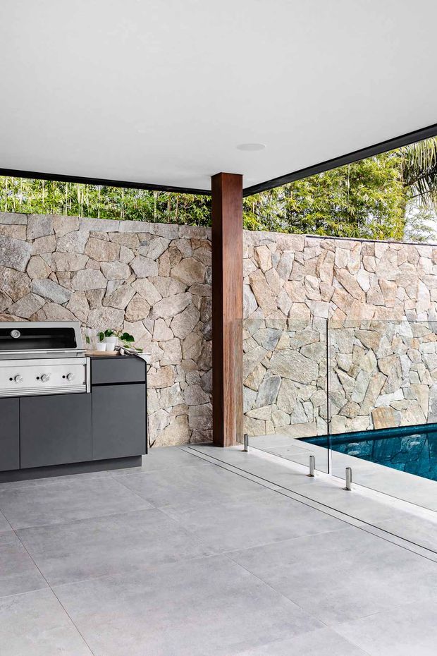 A contemporary covered outdoor entertaining area beside a swimming pool, with a built-in stainless-steel barbecue and dark cabinetry, large-format grey tiled paving, a warm timber support post, frameless glass pool fencing, and a rugged natural stone boundary wall. Dense planting is visible above the wall.