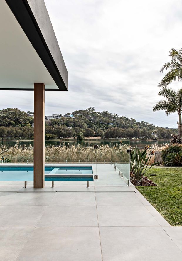 A contemporary outdoor terrace beside a swimming pool, overlooking a calm lake or waterway and densely vegetated opposite shore. The space features large pale concrete-look paving, frameless glass pool fencing, a slim flat roof with dark fascia, and a substantial timber support column. Tropical and native-style planting borders a lawn at right, creating a refined, tranquil waterfront setting.