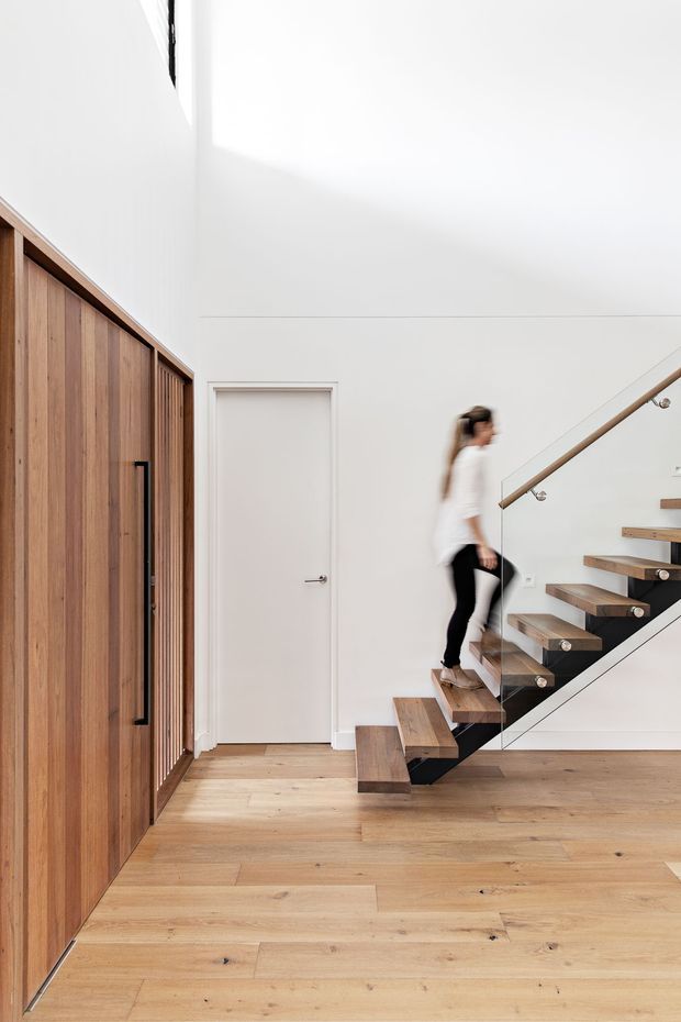 Bright minimalist interior hallway/entry space featuring a floating timber staircase with glass balustrade and slim metal handrail, a large built-in timber cupboard along the left wall, white walls and door, and wide-plank timber flooring. A person is ascending the stairs.