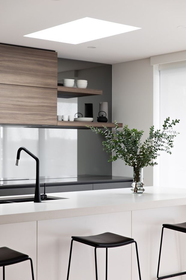 Bright contemporary kitchen featuring a large white island with integrated cabinetry and seating, a matte black gooseneck faucet, dark grey benchtop surfaces, warm wood-grain overhead cabinetry and open shelving, and a dark grey splashback wall. A large square skylight or flush ceiling light and sheer window covering provide soft natural illumination. Black minimalist stools and a glass vase with leafy branches add contrast.