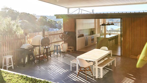 Sunlit outdoor entertaining area with a covered patio, dining table and chairs, barbecue grills, and a compact kitchen opening onto the deck. Vertical timber screening, a corrugated roof, and views toward greenery create a relaxed indoor-outdoor setting.