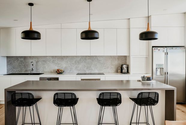 Contemporary minimalist kitchen with a large light-toned island, four black wire bar stools, flat-panel white cabinetry, patterned grey backsplash, stainless-steel appliances, and three oversized black pendant lights.