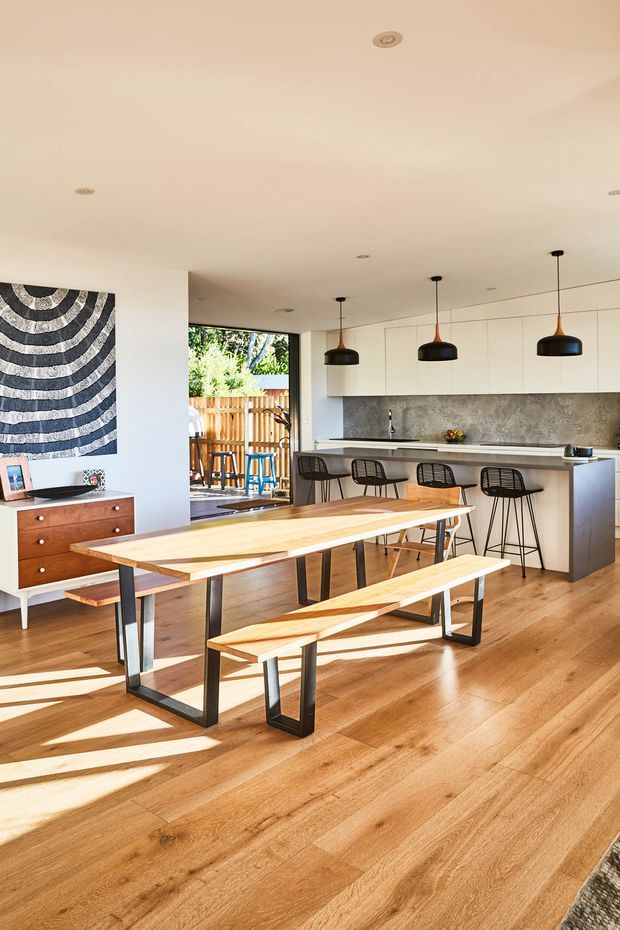 Bright open-plan kitchen and dining area with warm timber flooring, a long timber dining table and benches on black metal frames, a white kitchen with extensive upper cabinetry, stone-textured splashback, island breakfast bar, black stools, and three pendant lights. Large glazing opens toward a fenced outdoor area, while bold monochrome wall art and a timber sideboard add contrast.